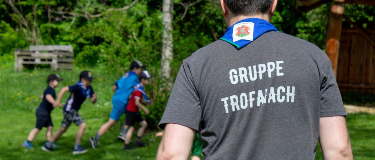 Ein Mann mit einem blauen Schal und einem grauen Shirt mit den Worten 'Gruppe Trofnach' auf dem Rücken steht auf einer Wiese.