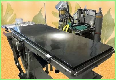 Ein schwarzer Operationstisch steht auf einem orangefarbenen Hintergrund, umgeben von medizinischen Geräten. Der Tisch ist glänzend und hat Räder. Verschiedene medizinische Werkzeuge und Geräte sind darum herum angeordnet.