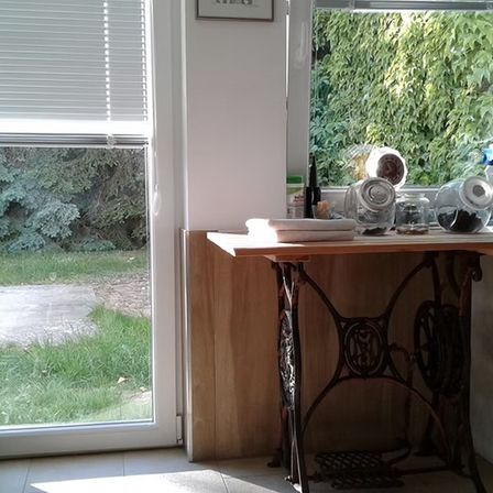 Eine Küchenecke mit einer Vintage-Nähmaschine unter einem Fenster, das einen Blick auf eine Rasenfläche draußen bietet. Der Tisch hat ein gefaltetes Handtuch und verschiedene Gläser.