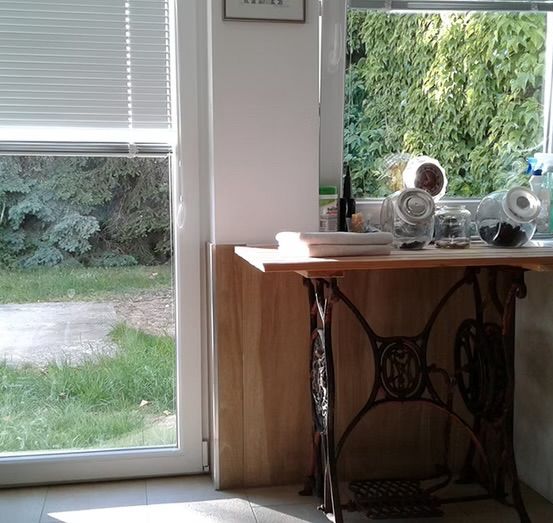 Eine Küchenecke mit einer Vintage-Nähmaschine unter einem Fenster, das einen Blick auf eine Rasenfläche draußen bietet. Der Tisch hat ein gefaltetes Handtuch und verschiedene Gläser.