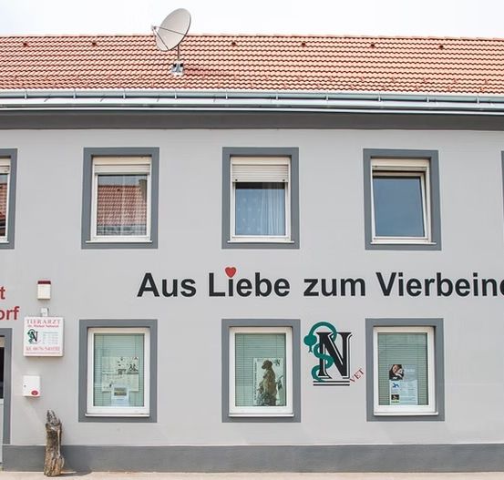 Das Gebäude hat ein rotes Dach, eine Satellitenschüssel und zeigt 'Aus Liebe zum Vierbeiner' mit einem Herz. Unten sind Fenster, eine Tür und ein 'Tierarzt Schwadorf'-Schild. Ein Schild auf der linken Seite zeigt 'Tierarzt' und Kontaktdaten.