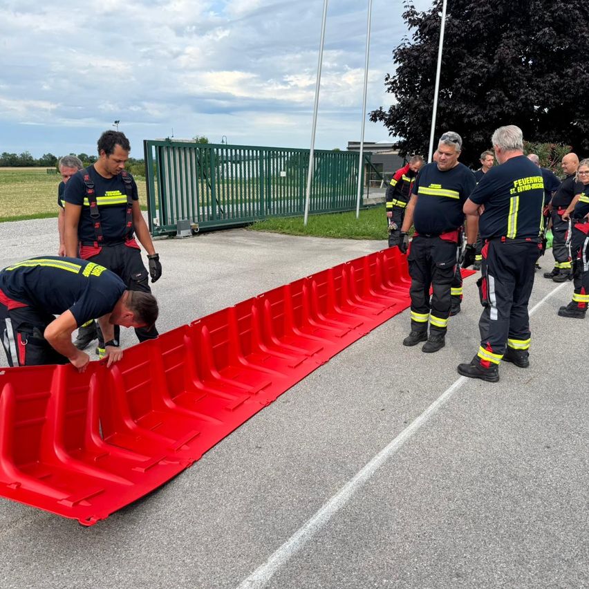 Eine Gruppe von Feuerwehrleuten übt das Aufstellen einer roten Barriere auf der Straße, während andere zusehen und ein grünes Tor dahinter steht.