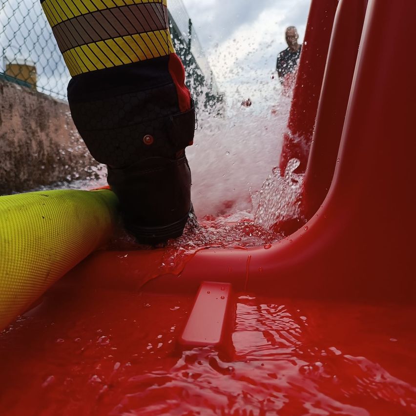 Ein Feuerwehrmann rutscht eine rote Wasserrutsche hinunter, neben einem gelben Schlauch, unter einem bewölkten Himmel.