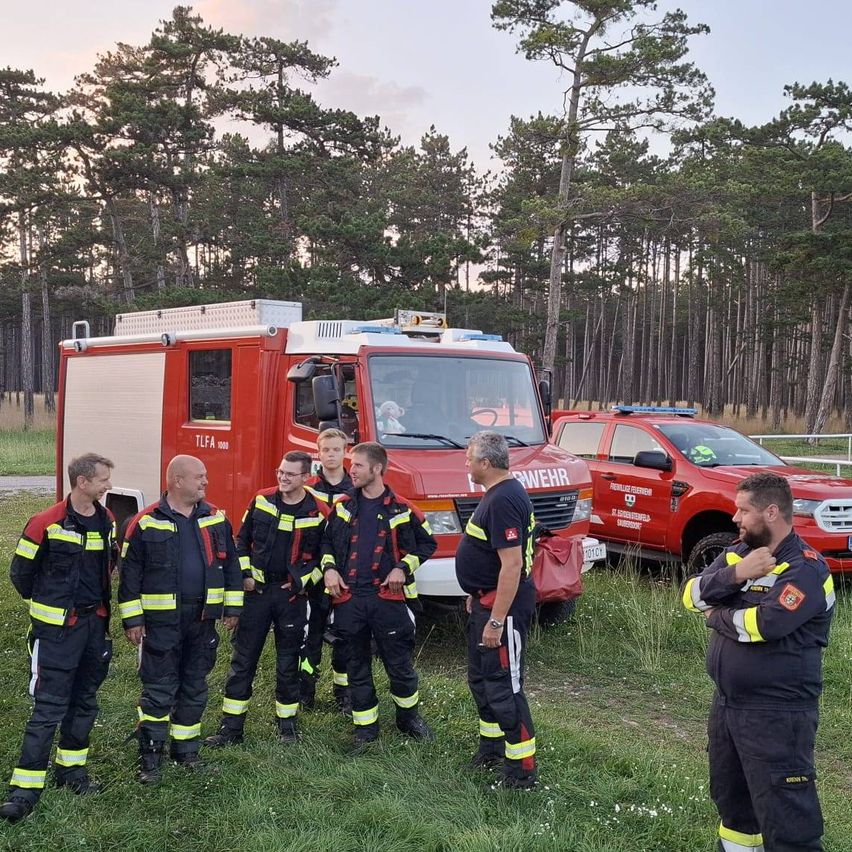 Eine Gruppe von Feuerwehrleuten steht auf einem Feld, mit zwei Feuerwehrwagen dahinter. Die Feuerwehrleute sind in voller Uniform gekleidet, und einer von ihnen hält eine rote Tasche.