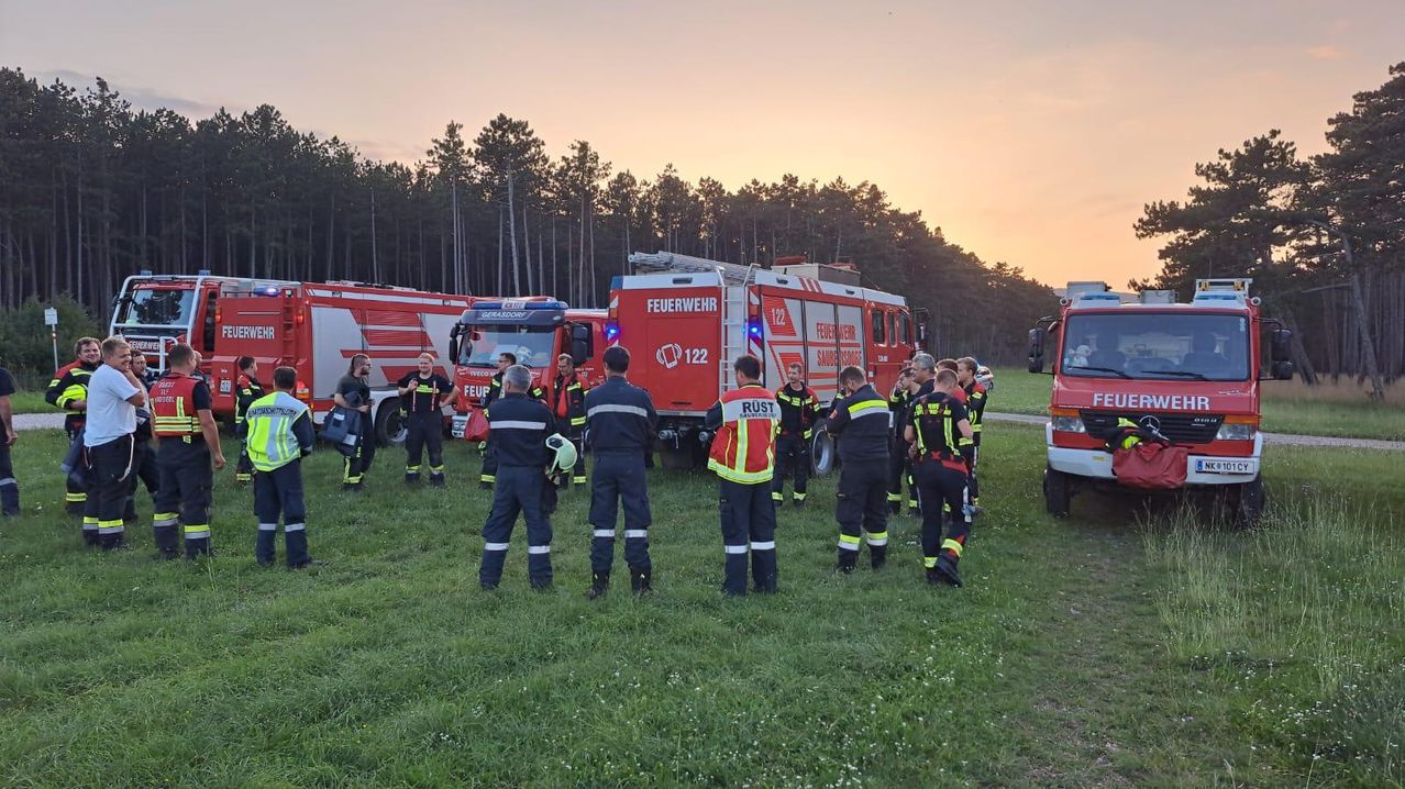 Eine Gruppe von Feuerwehrleuten steht auf einem Feld, mit geparkten Feuerwehrwagen dahinter. Der Himmel zeigt eine Mischung aus orange und rosa Tönen, was auf den Sonnenuntergang hinweist. Die Feuerwehrleute tragen Uniformen, einige von ihnen tragen Helme.
