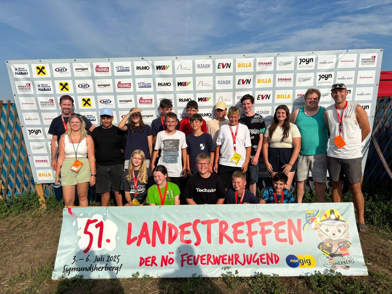 Eine Gruppe von Menschen, einschließlich Kindern, steht vor einer Wand mit Logos und trägt Ausweisbänder. Sie lächeln und posieren für ein Foto. Ein Banner trägt den Text '51. Landestreffen der No Feurwehrjugend'.