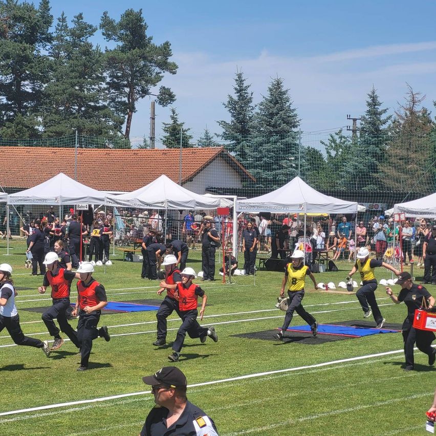 Eine Gruppe von Menschen in Uniformen läuft auf einem Feld mit Zelten und einer Menge von Zuschauern. Bäume und ein Gebäude mit einem roten Dach sind im Hintergrund.