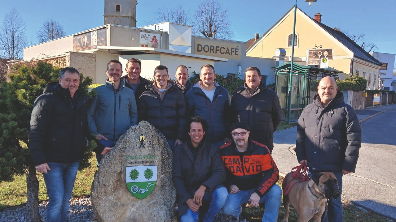 Eine Gruppe von Männern steht vor einem Stein mit einem Wappenschild in der Nähe eines Gebäudes mit dem Namen Dorfcafe. Die Männer tragen Winterkleidung.