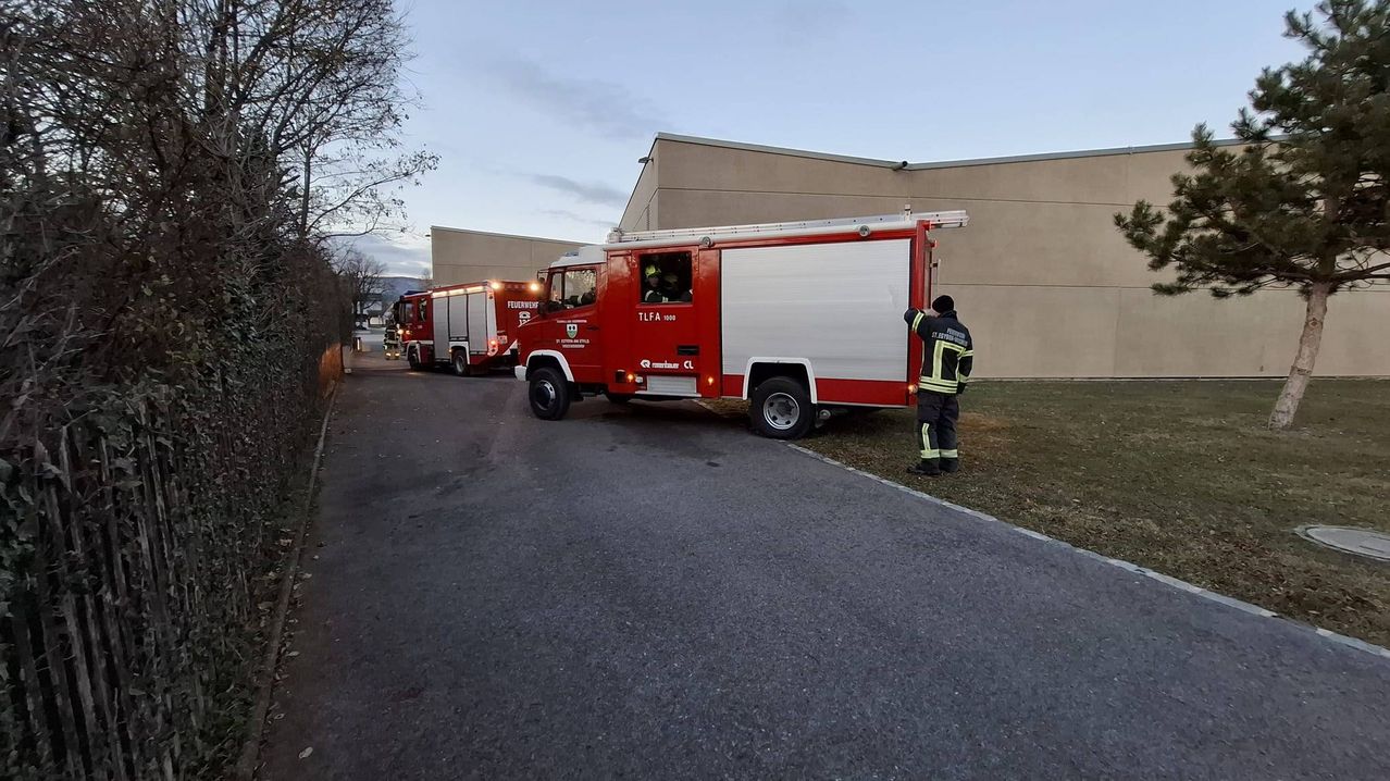 Zwei Feuerwehrfahrzeuge sind auf einer Straße geparkt, mit einem Gebäude im Hintergrund. Ein Feuerwehrmann steht neben einem der Fahrzeuge.
