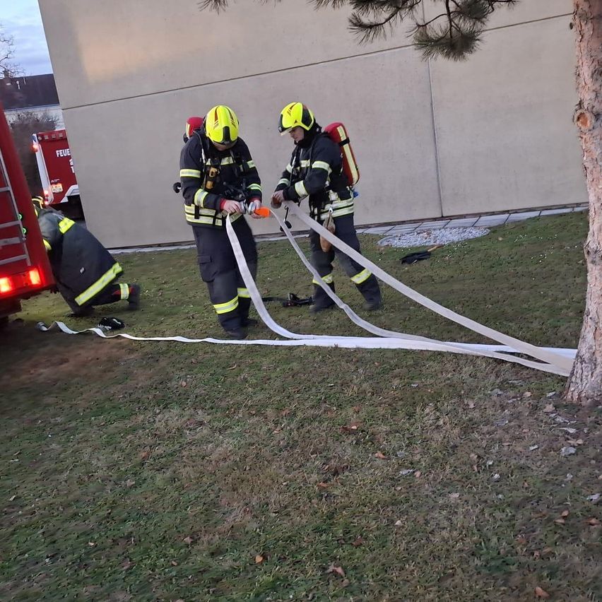 Drei Feuerwehrleute sind draußen und arbeiten mit Schläuchen. Einer kniet auf dem Boden, während die anderen stehen und die Schläuche halten. Ein Feuerwehrwagen ist in der Nähe geparkt.