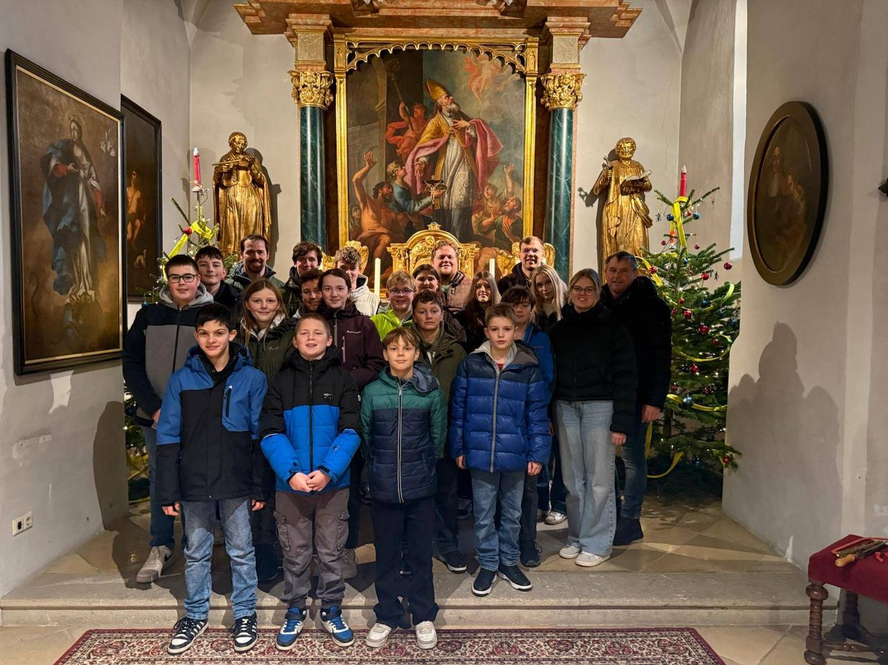 Eine Gruppe junger Menschen, Erwachsener und Kinder posiert für ein Foto in einer Kirche mit goldenen Statuen, einem großen Gemälde und einem Weihnachtsbaum im Hintergrund.