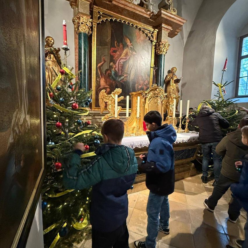 Mehrere Kinder schmücken einen Weihnachtsbaum in einer Kirche. Dahinter steht ein goldener Altar. Der Altar hat ein Gemälde und Kerzen.