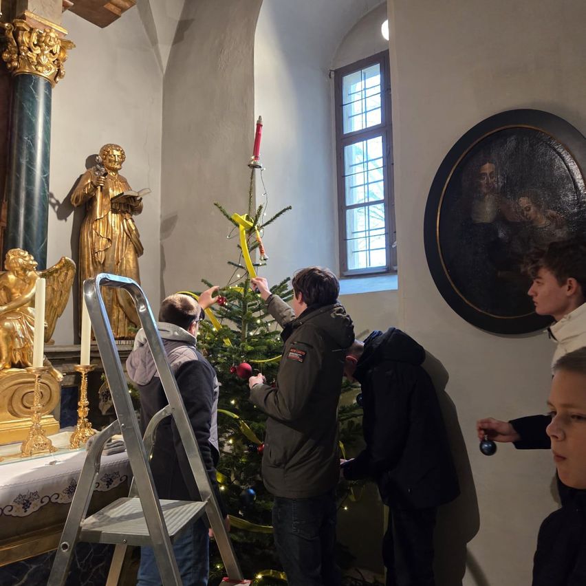 Mehrere Personen schmücken einen Weihnachtsbaum in einer Kirche. Eine goldene Statue und eine Kerze stehen auf dem Altar. Ein Gemälde hängt an der Wand.