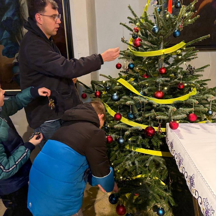 Drei Personen schmücken einen Weihnachtsbaum mit gelben Bändern und Ornamenten. Ein Mann leitet sie.
