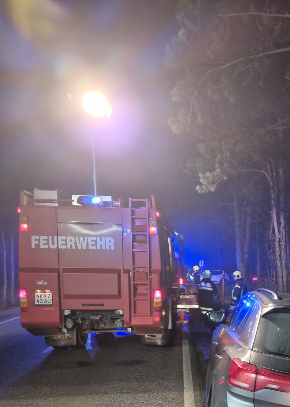 Ein Feuerwehrauto mit dem Wort Feuerwehr steht am Straßenrand. Es hat ein rotes und weißes Licht oben und eine Leiter hinten. Ein paar Leute stehen daneben.