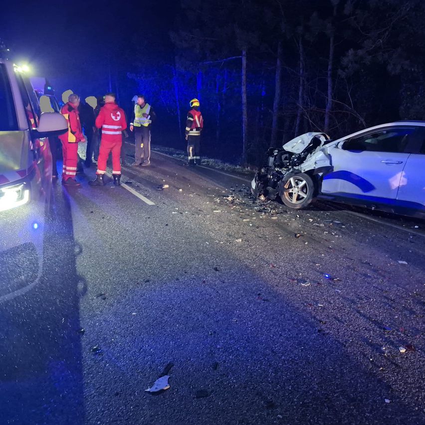 Rettungskräfte umgeben ein beschädigtes Auto auf einer dunklen Straße. Trümmer sind verstreut und Notbeleuchtung leuchtet im Hintergrund.