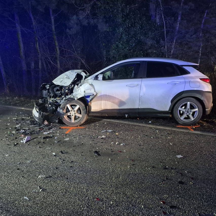 Ein weißer SUV ist nachts auf einer Straße verunglückt. Die Front ist schwer beschädigt, mit herumliegenden Trümmern. Bäume und ein dunkler Hintergrund sind sichtbar.
