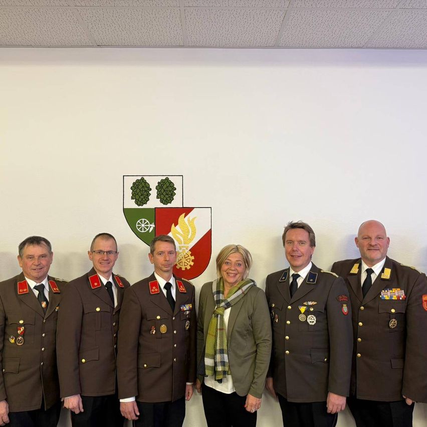 Eine Gruppe von Menschen in Uniformen, darunter eine Frau, steht zusammen vor einer Wand mit einem Wappen.
