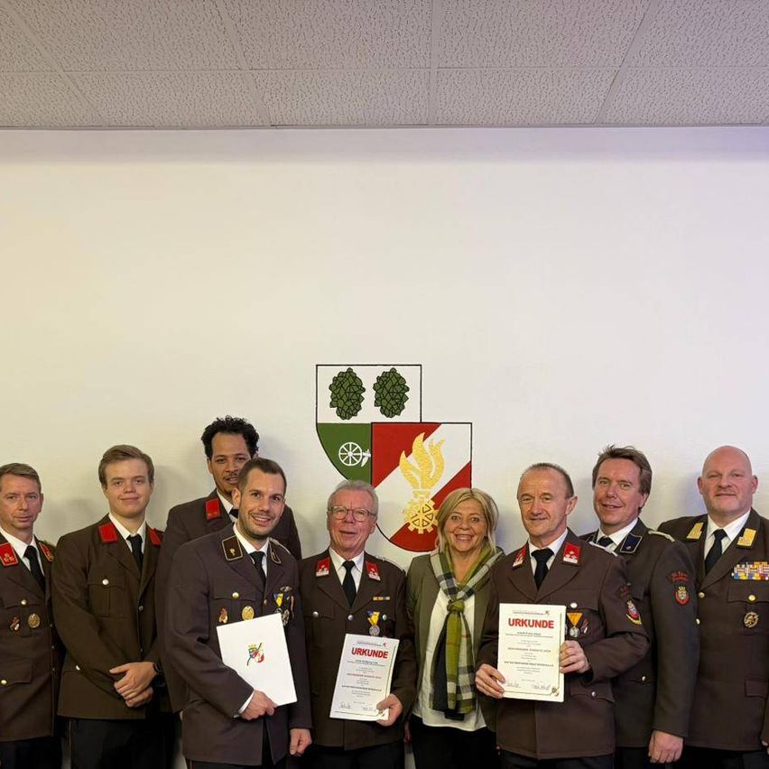 Eine Gruppe von Menschen in Militäruniformen, darunter eine Frau, steht zusammen vor einer Wand mit einem Logo. Sie halten Urkunden in den Händen.