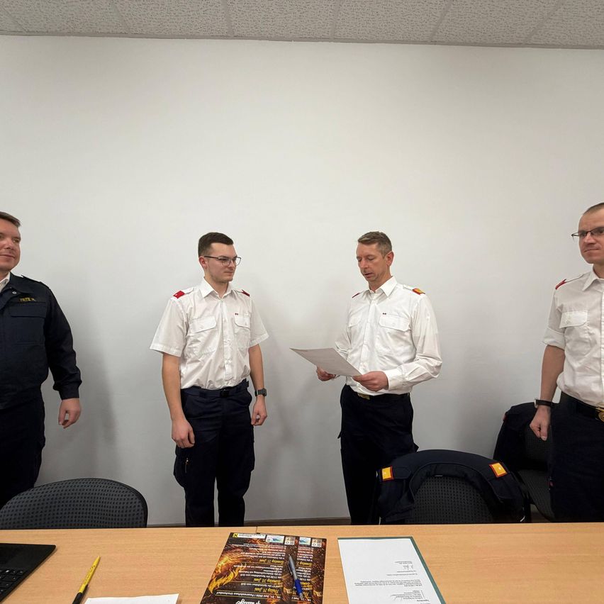 Four uniformed men stand in an office. One man reads a paper while others look on. Office chairs and a desk with a magazine and papers are in the background.