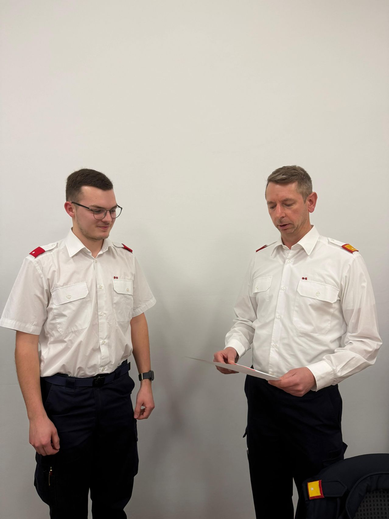 Two men in white uniforms are standing against a white wall, one holding a paper and looking at it. The other man is smiling.