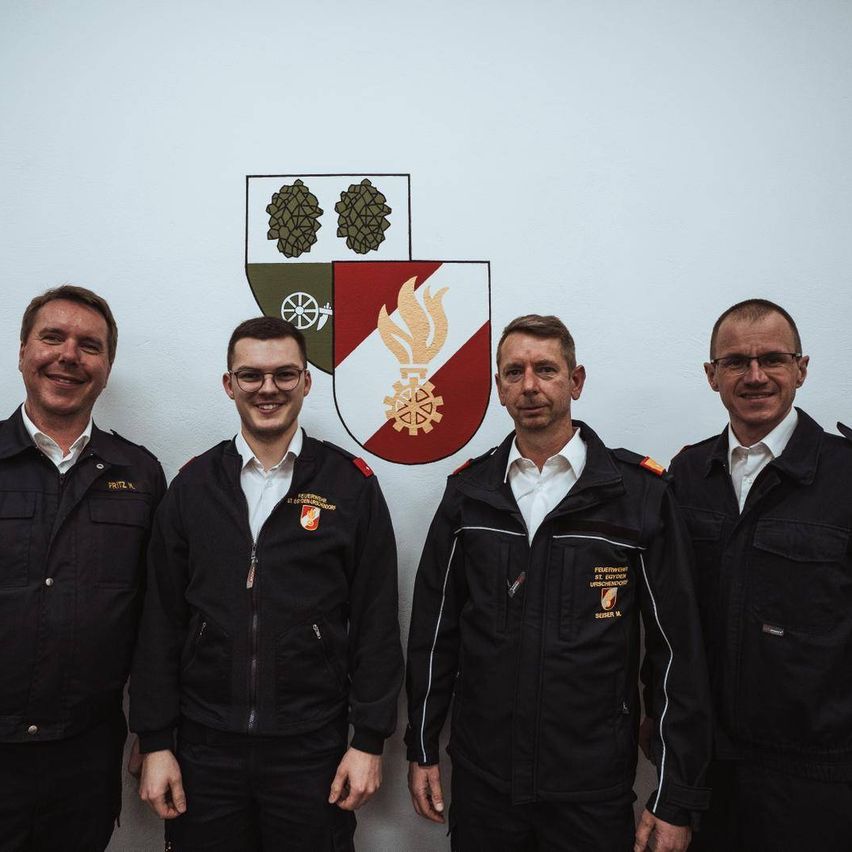 Four men in matching uniforms pose in front of a wall with a logo above them.