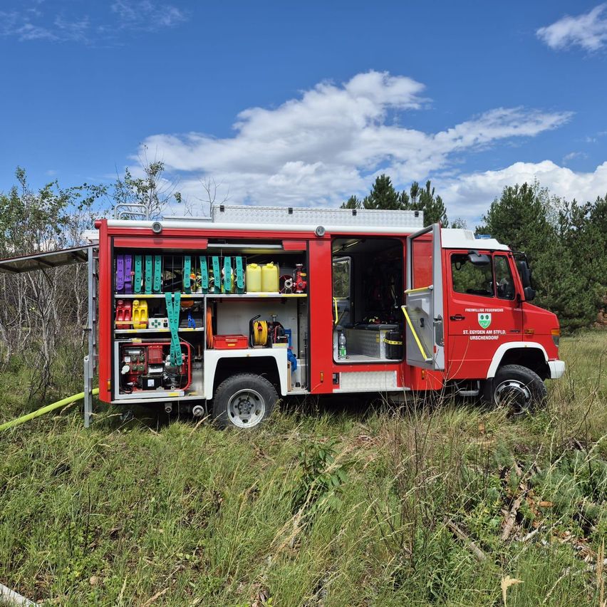 Ein roter Feuerwehrwagen mit offenen Türen, geparkt in einem Grasfeld mit Bäumen im Hintergrund.