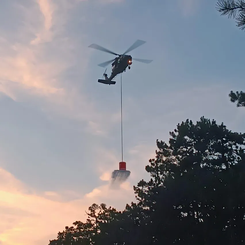 Ein Hubschrauber mit einem roten Eimer fliegt am Himmel und spritzt Wasser auf den Boden darunter. Der Himmel ist klar mit einigen Wolken.