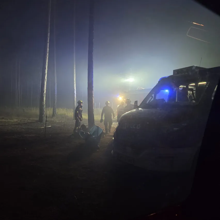 Eine Gruppe von Menschen mit Helmen steht in einem dunklen Bereich mit einem Fahrzeug vor ihnen. Der Bereich ist von Bäumen umgeben und von einem Licht beleuchtet.