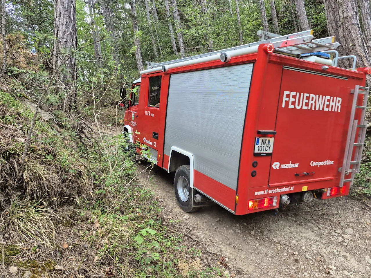 Ein roter Feuerwehrwagen steht auf einem Feldweg, umgeben von Bäumen und Büschen. Auf der Rückseite des Wagens steht das Wort 'FEUERWEHR'.