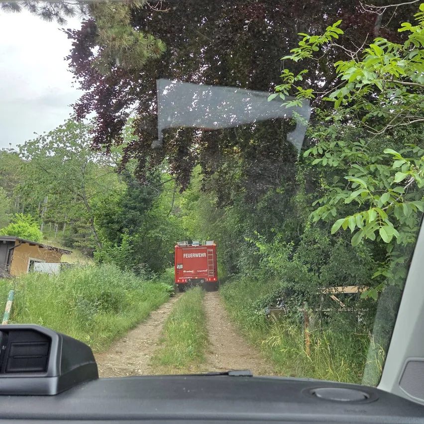 Ein roter Feuerwehrwagen fährt auf einem schmalen Feldweg zwischen Bäumen und Büschen. Der Weg verläuft zwischen einem Haus und einem Wald.