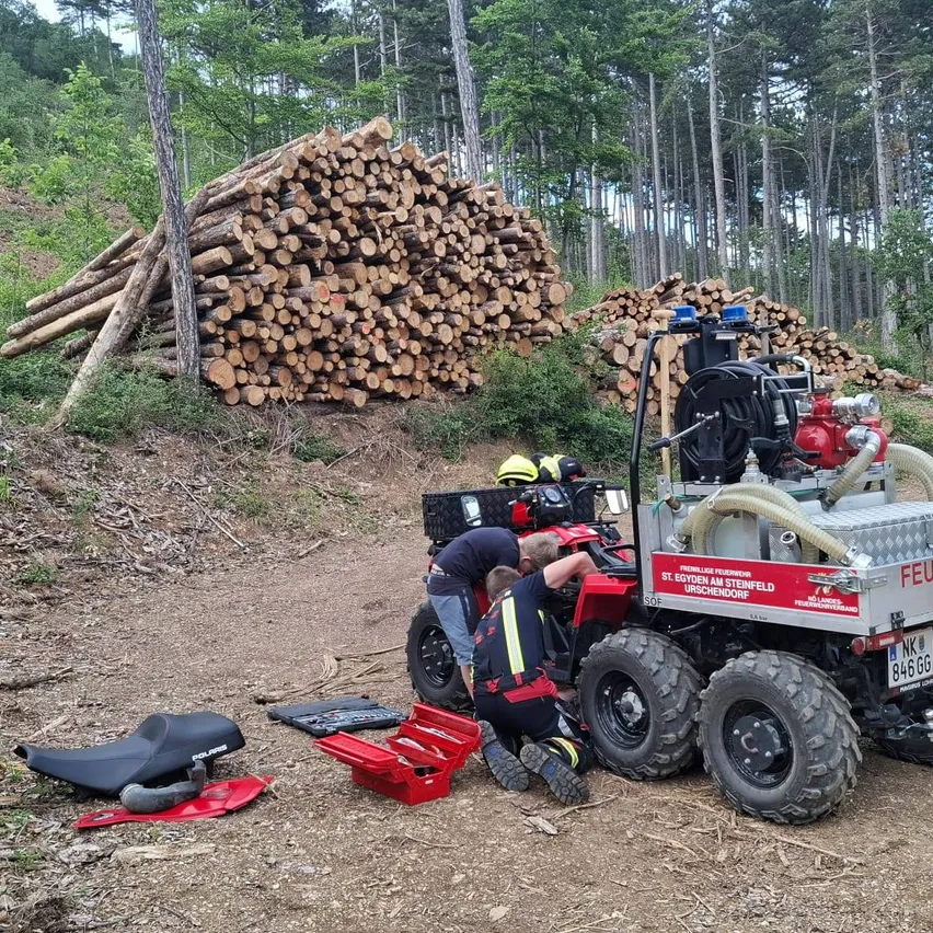 Zwei Feuerwehrleute arbeiten an einem Feuerwehr-Vierrad in einem Wald. Dahinter sind Stapel von Baumstämmen. Ein Werkzeugkasten und Werkzeuge liegen auf dem Boden.