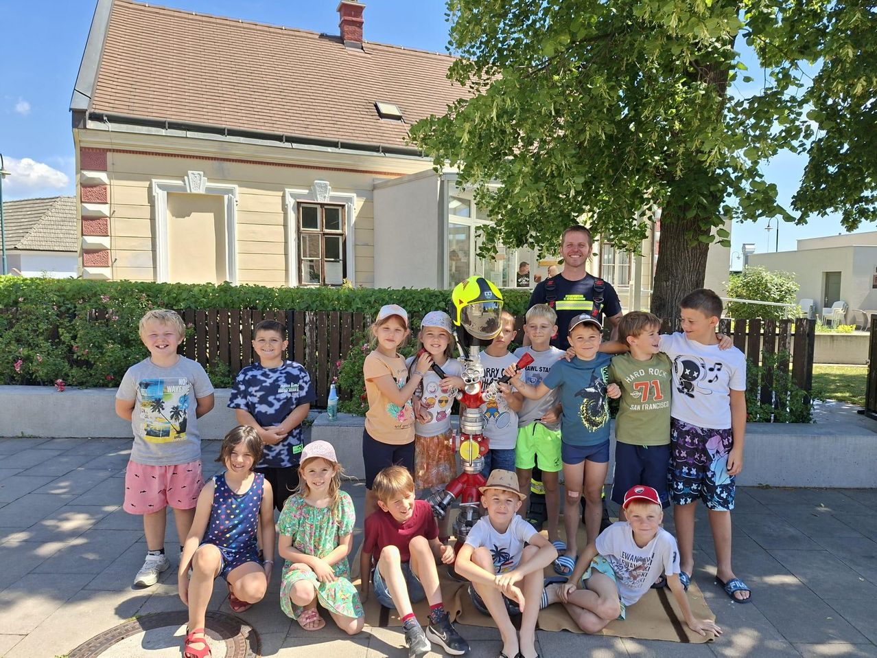 Eine Gruppe von Kindern und Erwachsenen, darunter ein Feuerwehrmann, posiert für ein Foto vor einem Baum und einem Haus.
