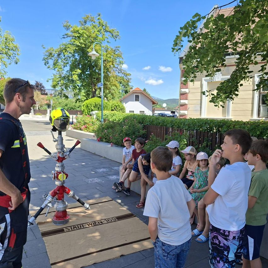 Ein Feuerwehrmann demonstriert eine Feuerwehrhydrant an eine Gruppe von Kindern, die auf einer Mauer sitzen.
