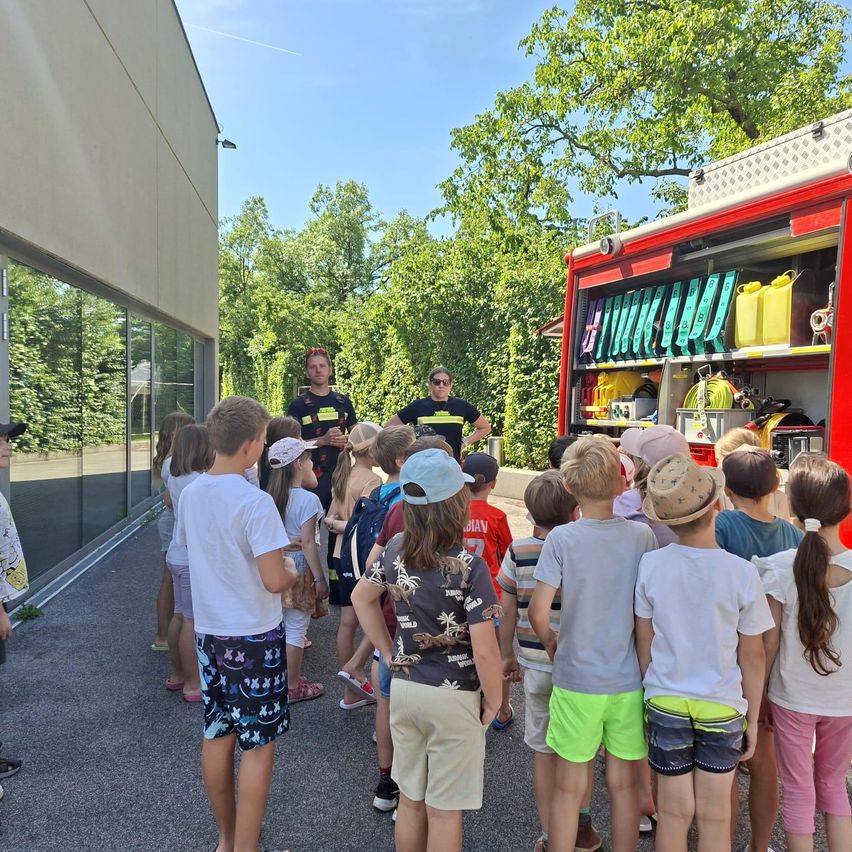 Kinder versammeln sich um einen Feuerwehrwagen, zwei Feuerwehrleute stehen hinten, einer zeigt Ausrüstung. Bäume und ein Gebäude im Hintergrund.