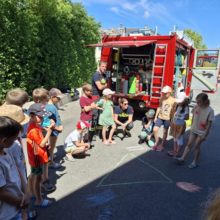 Ein Feuerwehrmann erklärt einer Gruppe von Kindern um ein Feuerwehrauto herum die Brandschutzmaßnahmen. Sie sind in einem Kreis versammelt mit Kreidezeichnungen auf dem Boden.