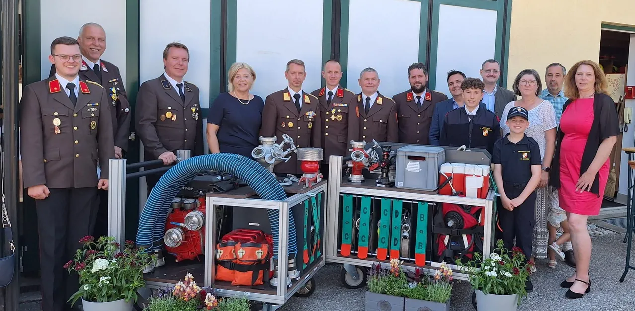 Eine Gruppe von Militäroffizieren und einer Frau steht neben Feuerwehrfahrzeugen. Sie tragen Uniformen und Medaillen. Auf den Trolleys befinden sich mehrere Gegenstände, darunter ein Feuerlöscher, ein Hydrant und eine Aufbewahrungsbox.