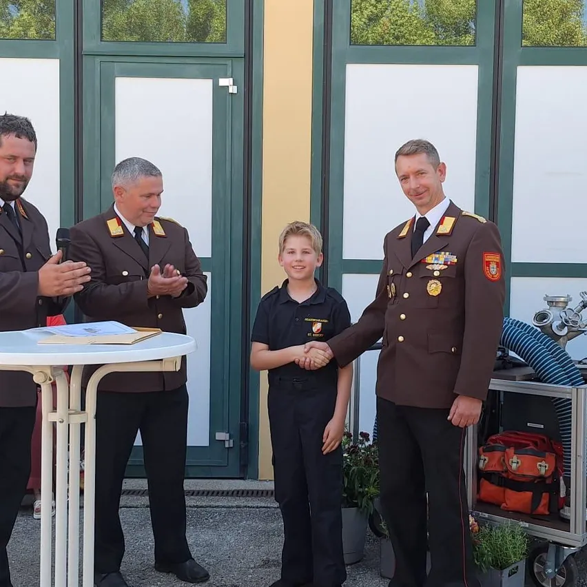 Drei Männer in Militäruniformen, einer mit einem Mikrofon, stehen neben einem Tisch. Ein Junge in einer schwarzen Uniform schüttelt die Hand eines Mannes. Sie stehen vor einem Gebäude mit Glastüren und einem Wagen mit Ausrüstung.