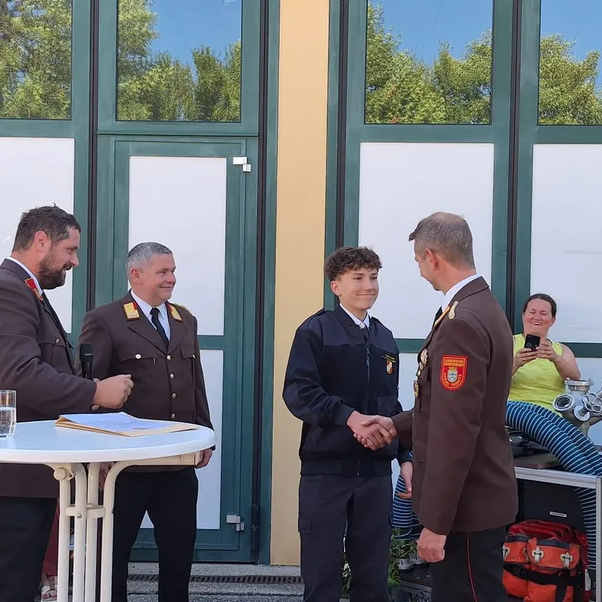 Ein junger Mann schüttelt die Hände von zwei älteren Männern in Uniform, die vor einem Gebäude mit Glastüren und Fenstern stehen. In der Nähe hält eine Frau ein Telefon.