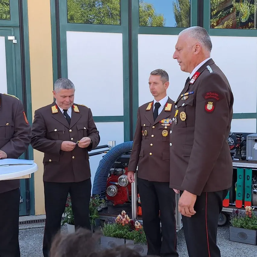 Drei Männer in braunen Uniformen stehen im Freien, einer mit zusammengefalteten Händen. Hinter ihnen steht ein Gebäude mit grünen Fenstern. Ein Feuerlöscher ist sichtbar.
