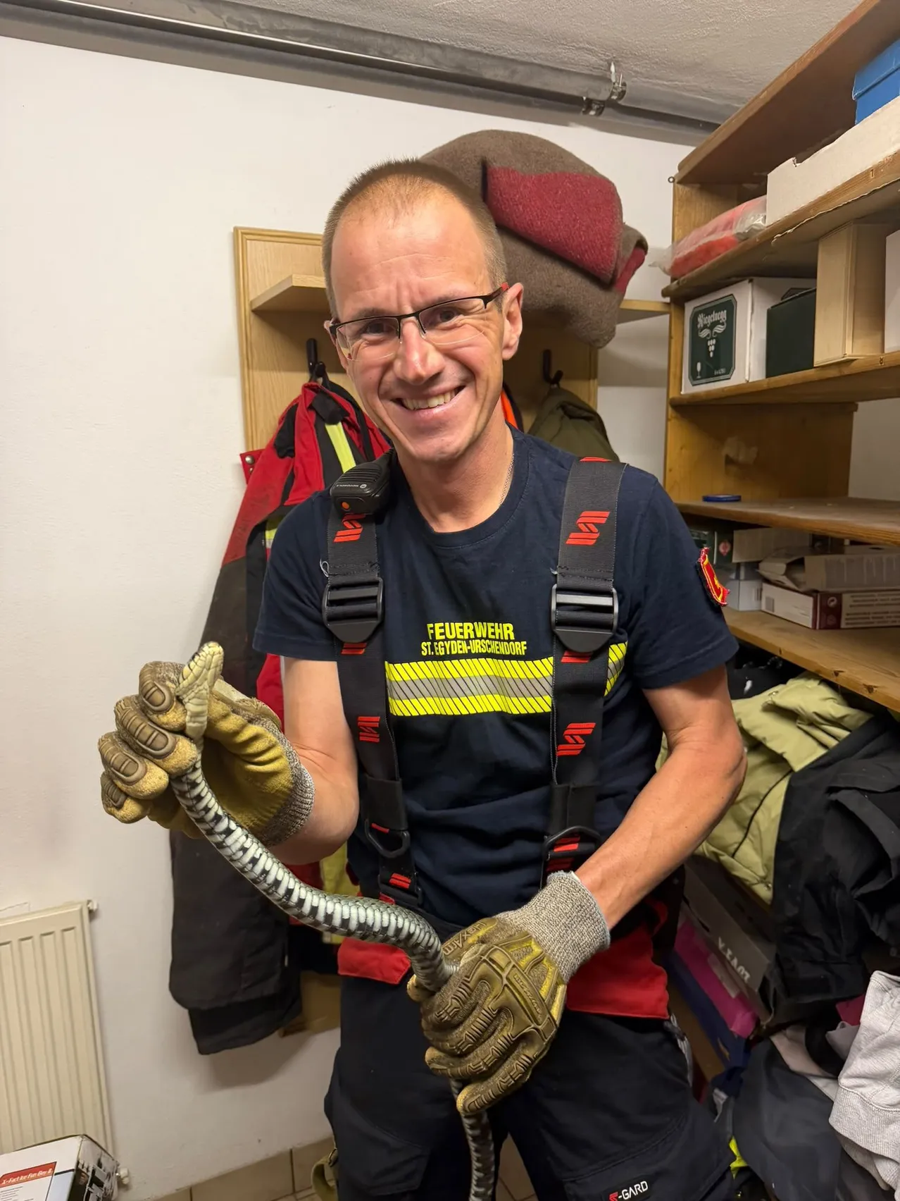 Ein Feuerwehrmann in Uniform hält eine aufgerollte Seilrolle und lächelt, mit Ausrüstung im Hintergrund.