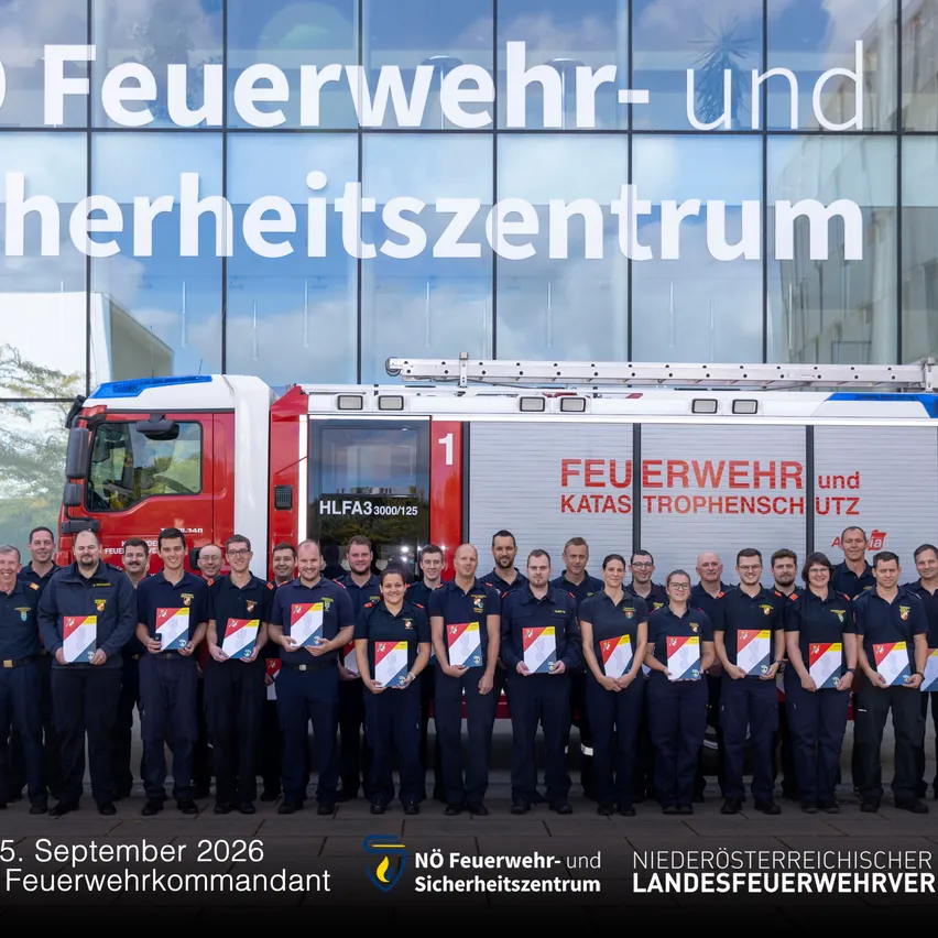 Eine Gruppe von Feuerwehrleuten in Uniform steht vor einem Feuerwehrwagen, mit einem Gebäude im Hintergrund. Sie halten Urkunden in den Händen und posieren für ein Foto.