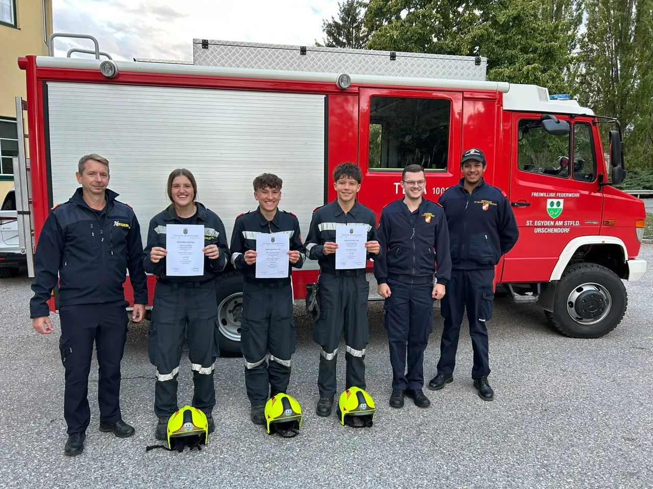Fünf Feuerwehrleute stehen vor einem roten Feuerwehrauto. Sie tragen alle Uniformen und halten Urkunden in den Händen.