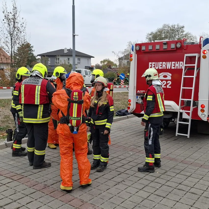 Eine Gruppe von Feuerwehrleuten in Schutzkleidung steht vor einem roten Feuerwehrwagen auf einem gepflasterten Bürgersteig.