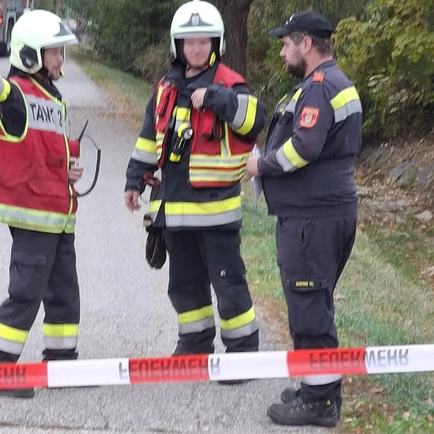 Drei Feuerwehrleute stehen auf einer Straße, einer hält eine Seilbarriere. Sie sind in voller Feuerwehrausrüstung gekleidet, einschließlich Helmen und Westen.