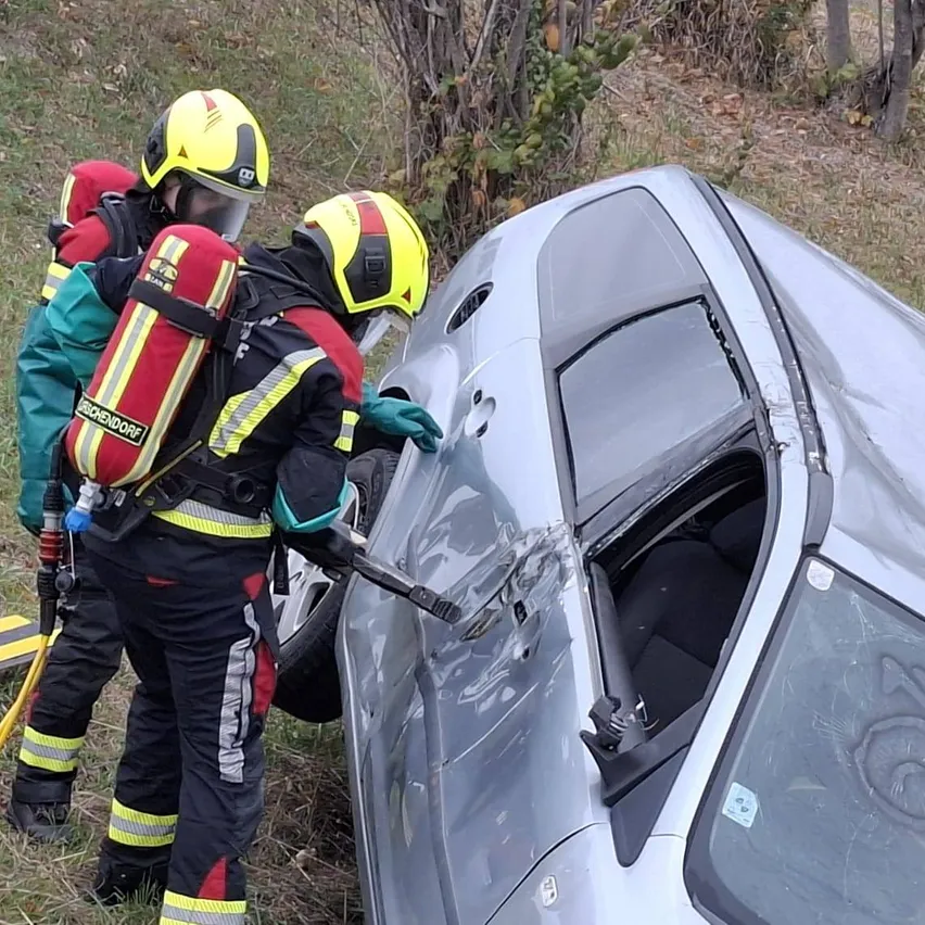 Zwei Feuerwehrleute in voller Ausrüstung arbeiten an einem umgekippten Auto. Einer hält ein Werkzeug, der andere einen Feuerlöscher.
