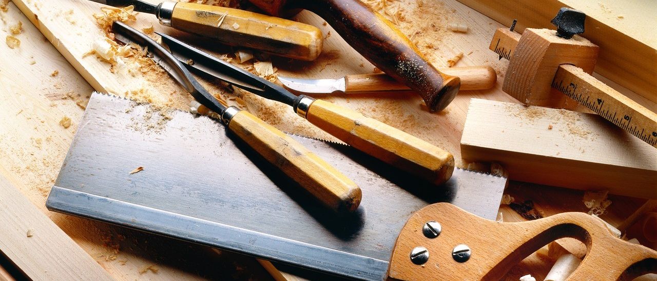 Ein Holzarbeitstisch zeigt verschiedene Werkzeuge, darunter eine Säge, Meißel und einen Hammer. Holzspäne sind auf der Oberfläche verstreut.