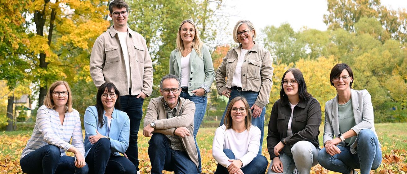 Eine Gruppe von Menschen posiert für ein Foto in einem Park mit Herbstblättern auf dem Boden. Sie tragen alle eine Brille und lächeln.