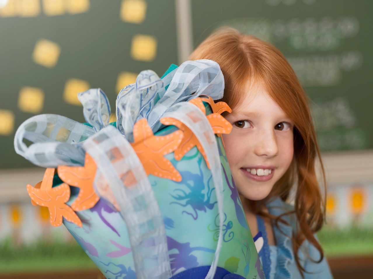 Ein junges Mädchen mit roten Haaren hält eine bunte Geschenktüte mit Seesternen und Schleifen. Sie lächelt und posiert für ein Foto im Klassenzimmer.