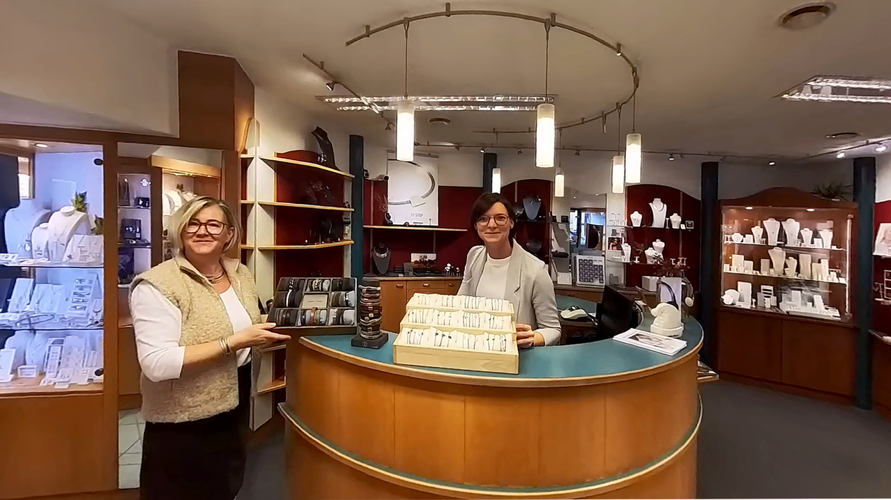 Zwei Frauen in einem Juwelierladen, eine hält eine Schmuckschatulle. Eine Frau hinter der Theke lächelt und schaut in die Kamera.